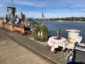 Memorial table with ship's bell and wreath on a Mare Island pier beside a docked naval ship